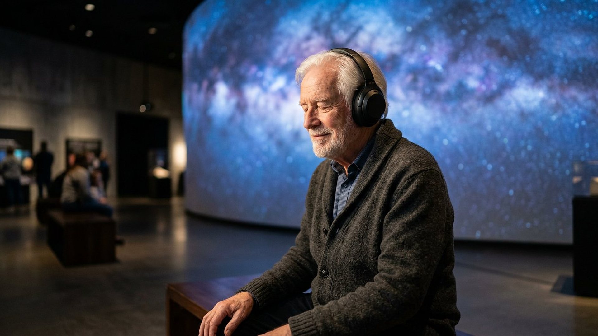 Un homme âgé aux cheveux blancs, casque audio sur les oreilles, écoute les yeux fermés en souriant, assis dans une salle de musée devant un grand écran courbe où brille la Voie lactée.