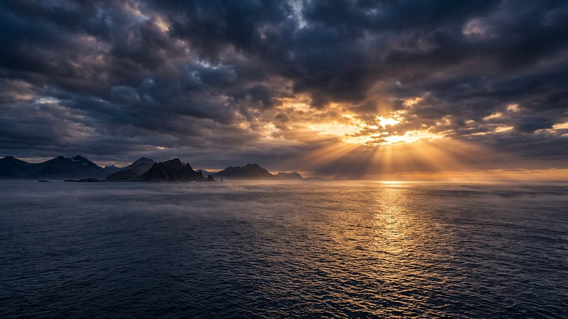 Le soleil perce une épaisse couche de nuages au-dessus d'un océan calme ; des rayons dorés descendent vers l'eau, des montagnes sombres à l'horizon.
